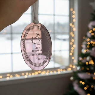 Decorative ornament with engraved text, held by a hand in front of a Christmas tree and window.