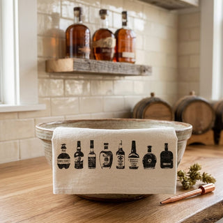 Bowl with a towel featuring whiskey bottle prints on a wooden surface, with barrels and bottles in the background.
