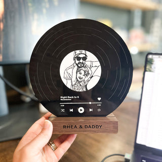 Round black record with a wooden base, held by a hand, featuring a photo and text.